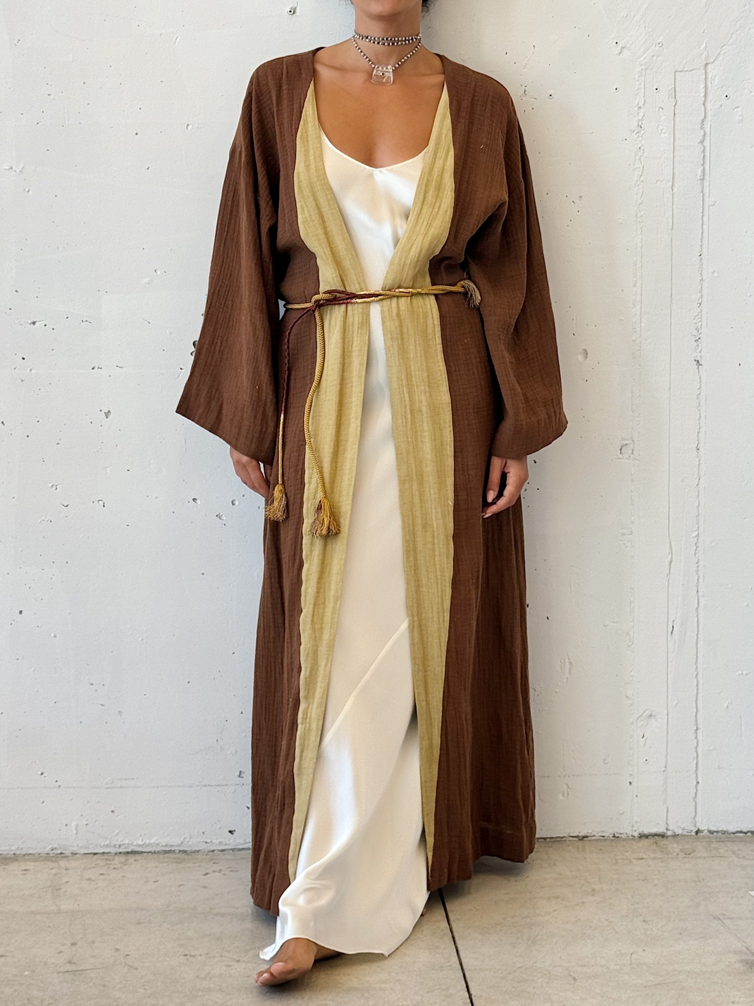 Woman wearing a long brown robe over a white dress against a white wall.