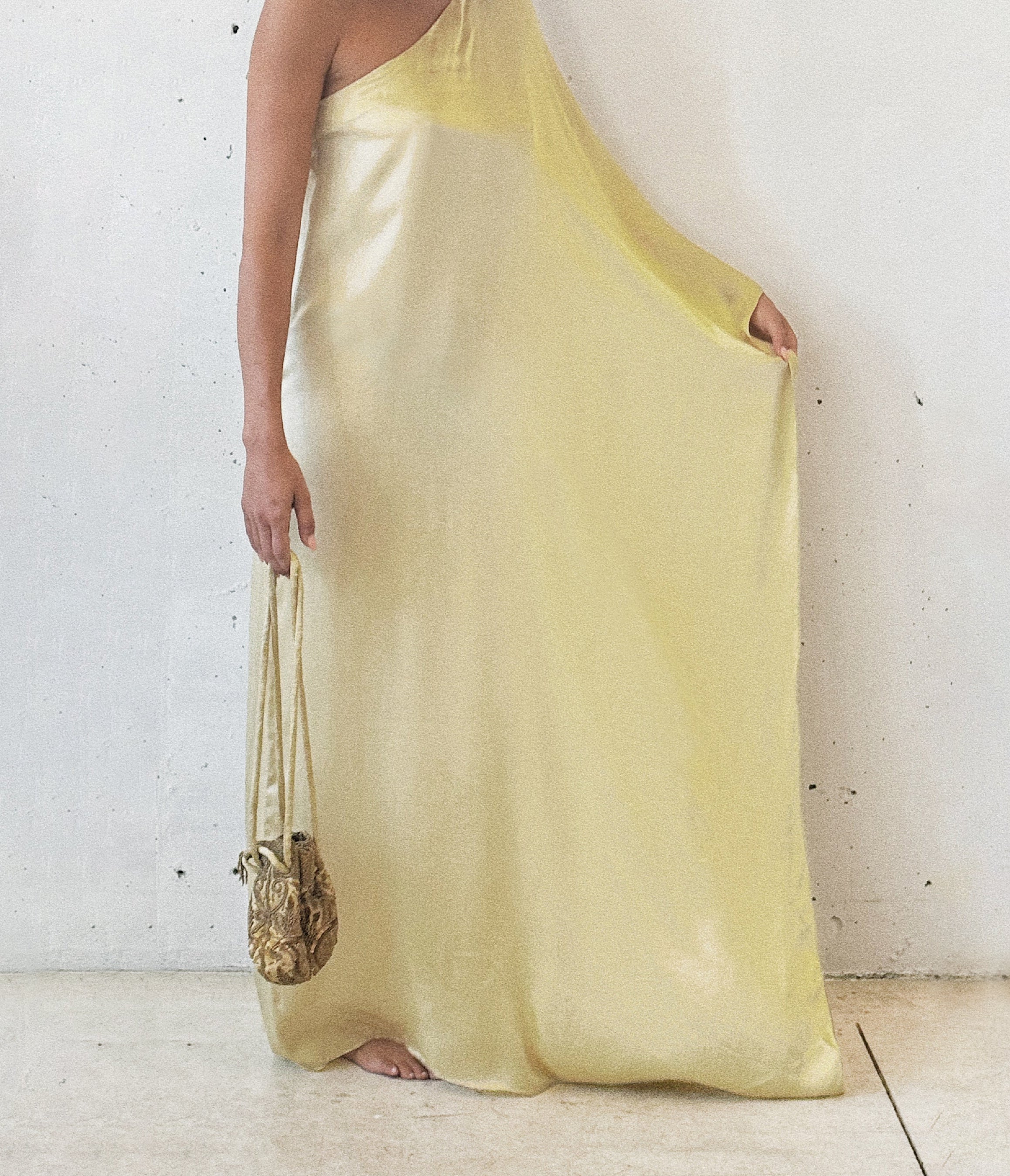 A person wearing a light yellow, organic silk, maxi dress with a matching scarf, standing against a white background, carrying a small gold bag.