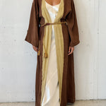 Woman wearing a long brown robe over a white dress against a white wall.
