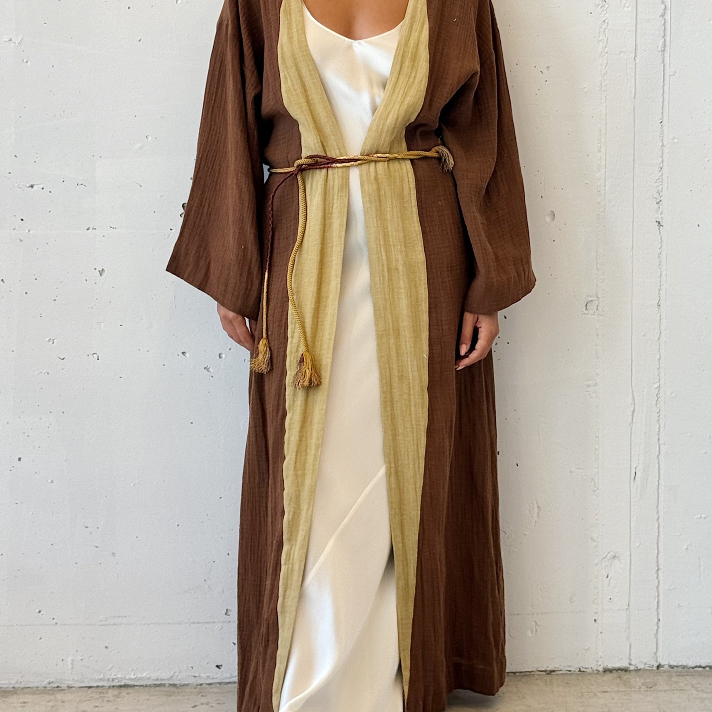 Woman wearing a long brown robe over a white dress against a white wall.