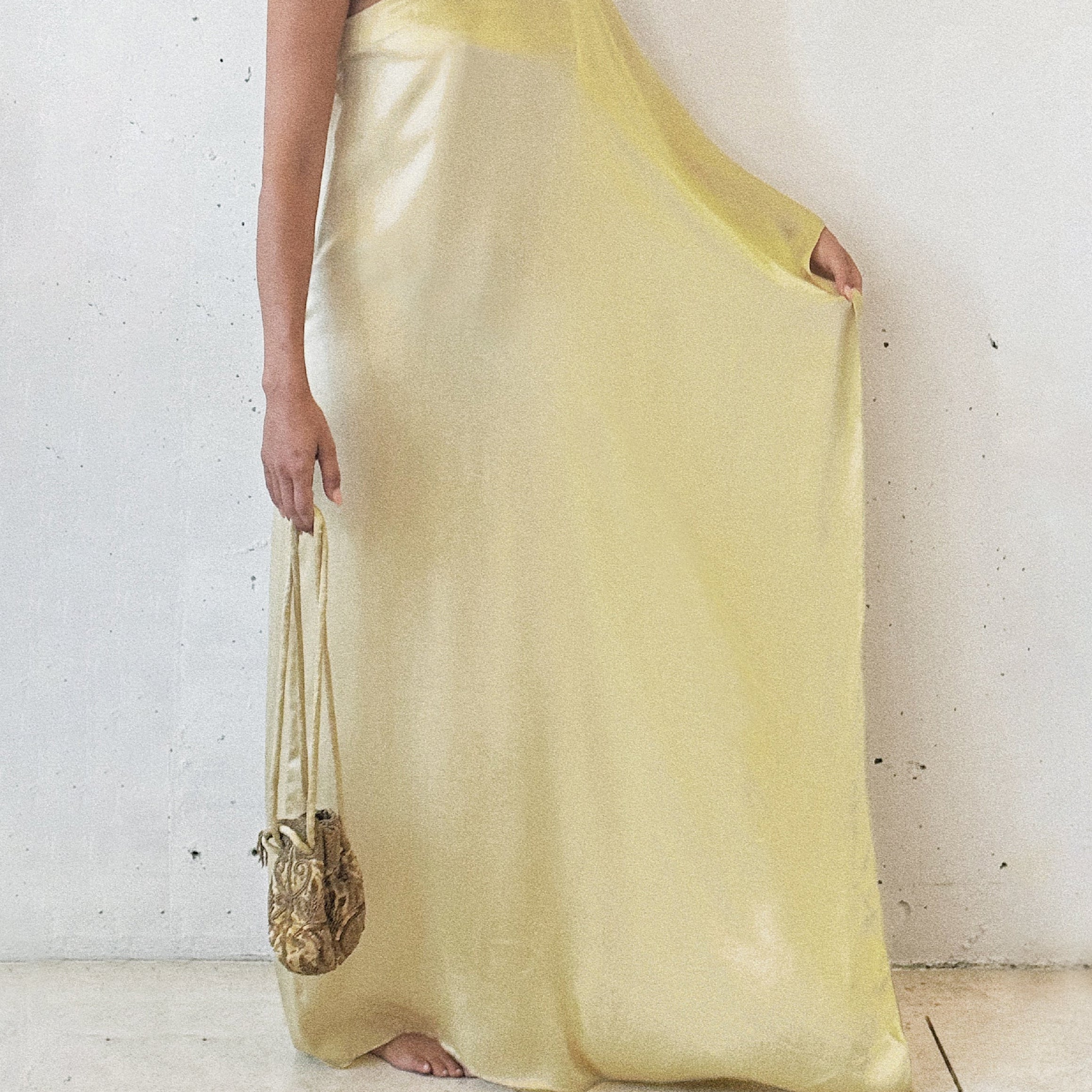 A person wearing a light yellow, organic silk, maxi dress with a matching scarf, standing against a white background, carrying a small gold bag.